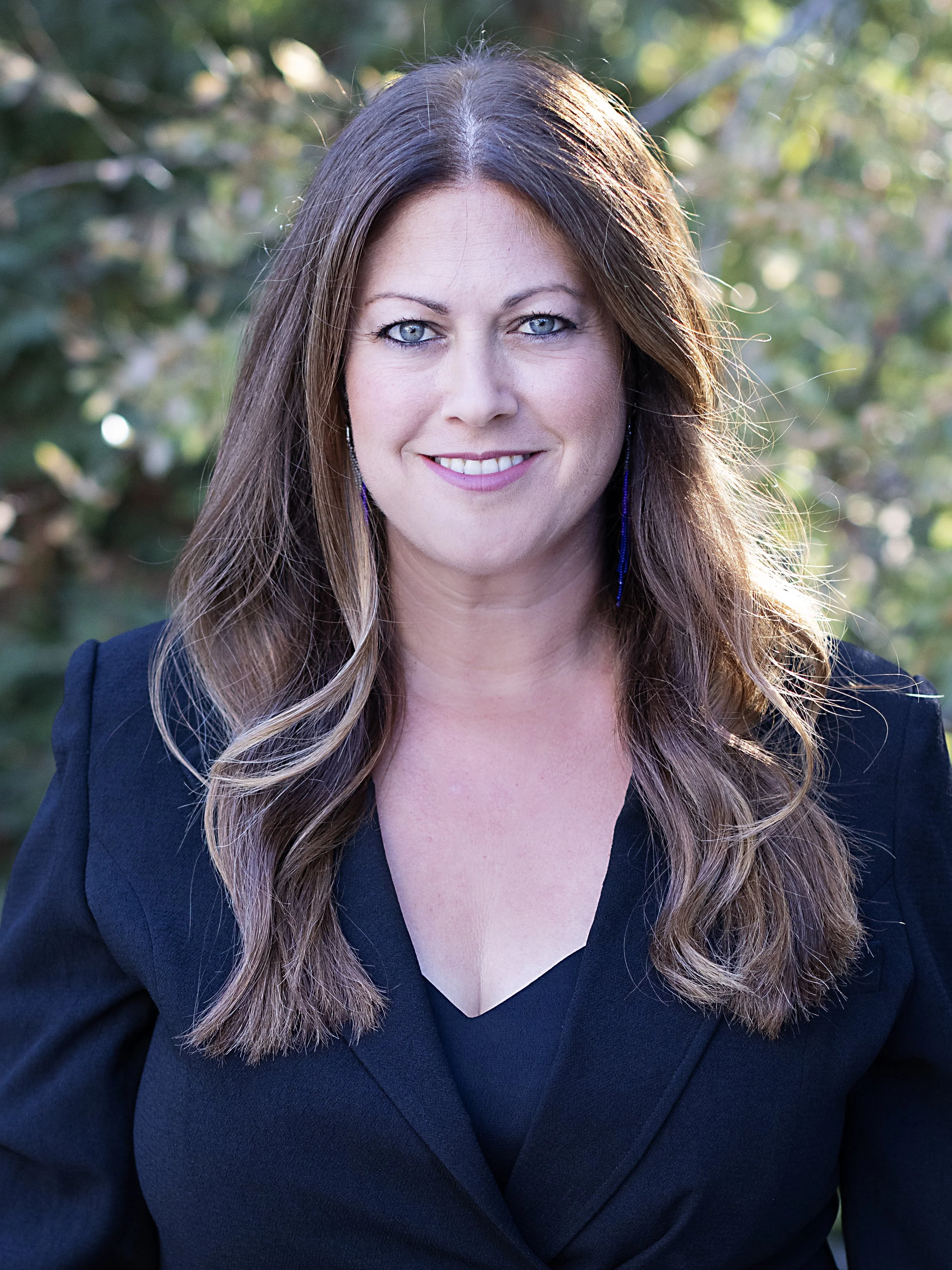 Close-up portrait of a woman with long wavy light brown hair, wearing a black blazer and purple earrings, smiling outdoors with blurred green foliage in the background.