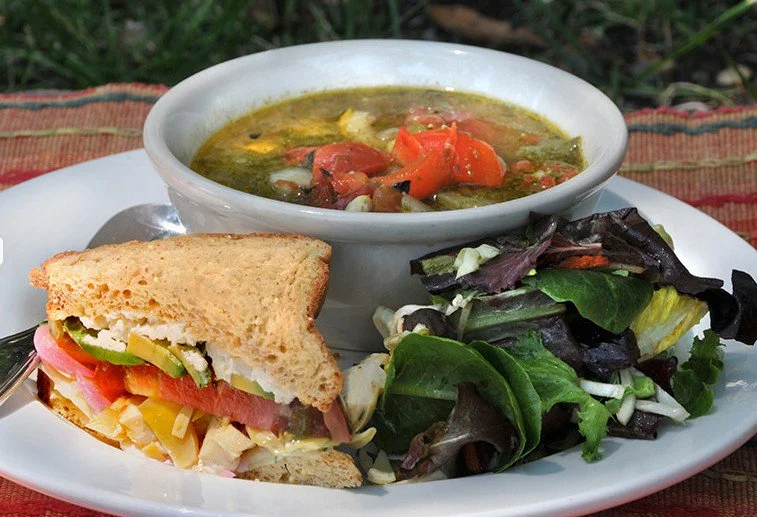 A plate with a bowl of vegetable soup, a half sandwich with tomato, lettuce, cheese, and turkey, and a side salad of mixed greens.