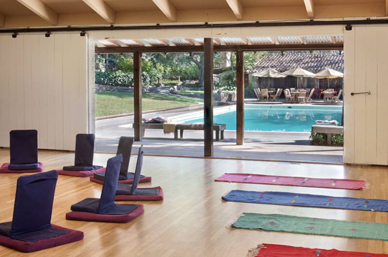 Indoor yoga room with mats and cushions, overlooking a patio and swimming pool outside.