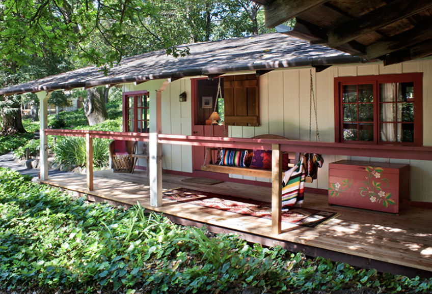 A cozy porch with a swing surrounded by lush green trees and plants, featuring red-trimmed windows and a floral-painted pink chest.
