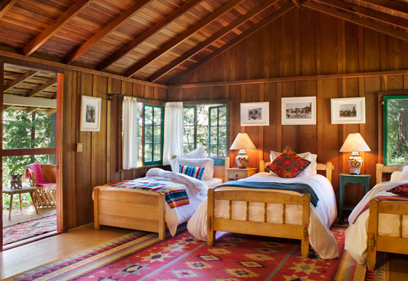 A rustic wooden bedroom with three beds, colorful bedding, and two lamps on nightstands. Open windows reveal a green outdoor view. A door leads to a porch with outdoor chairs and a dog.