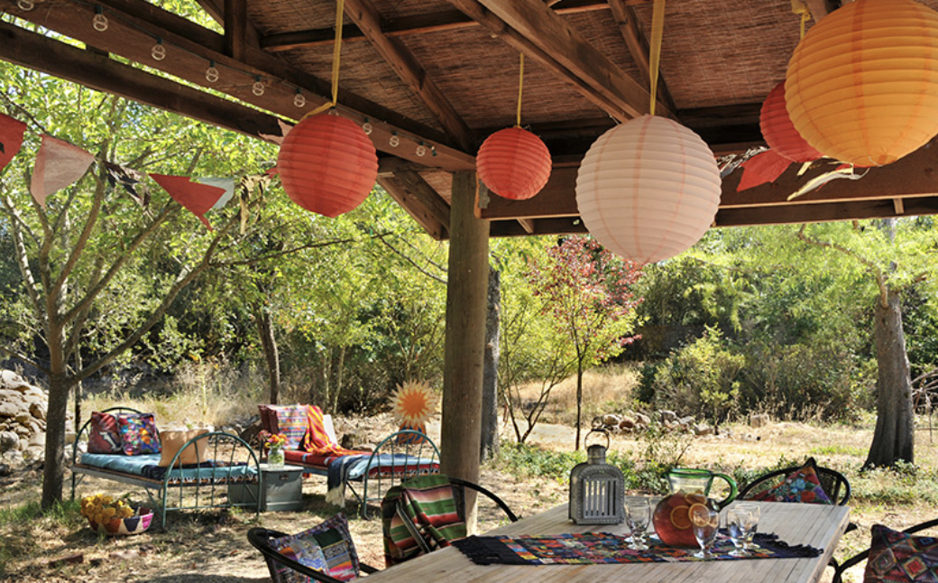 Outdoor patio decorated with colorful paper lanterns, bunting, and colorful cushions on metal chairs and outdoor sofas, surrounded by trees and natural scenery.