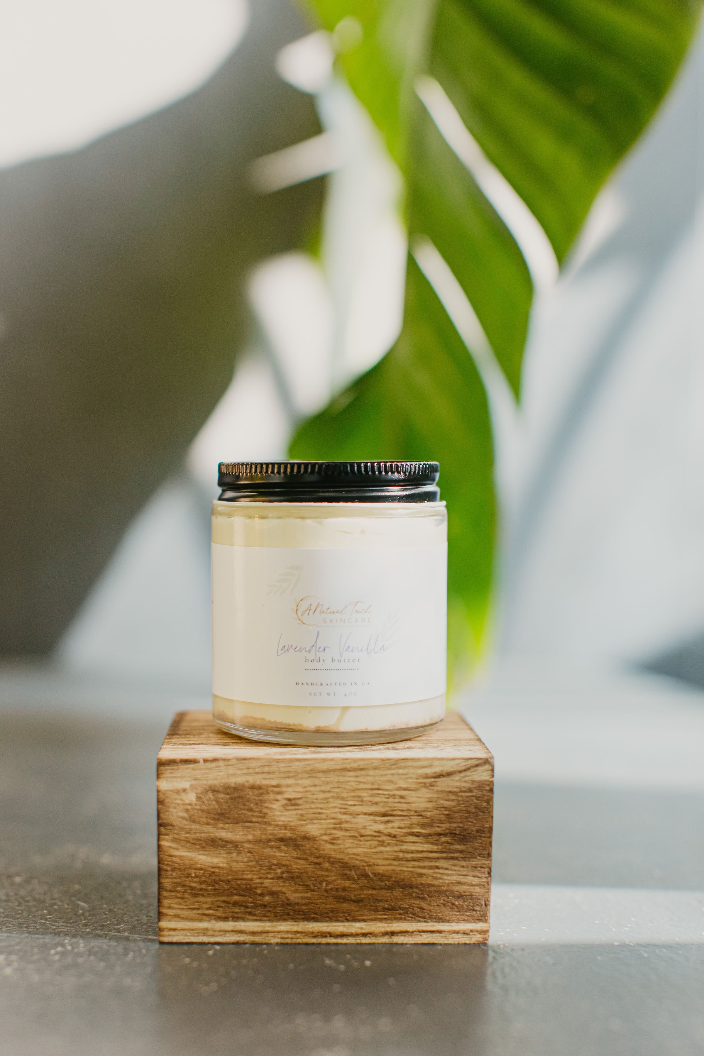 A jar of lavender vanilla body butter with a black lid, placed on a wooden block with a green leafy plant in the background.