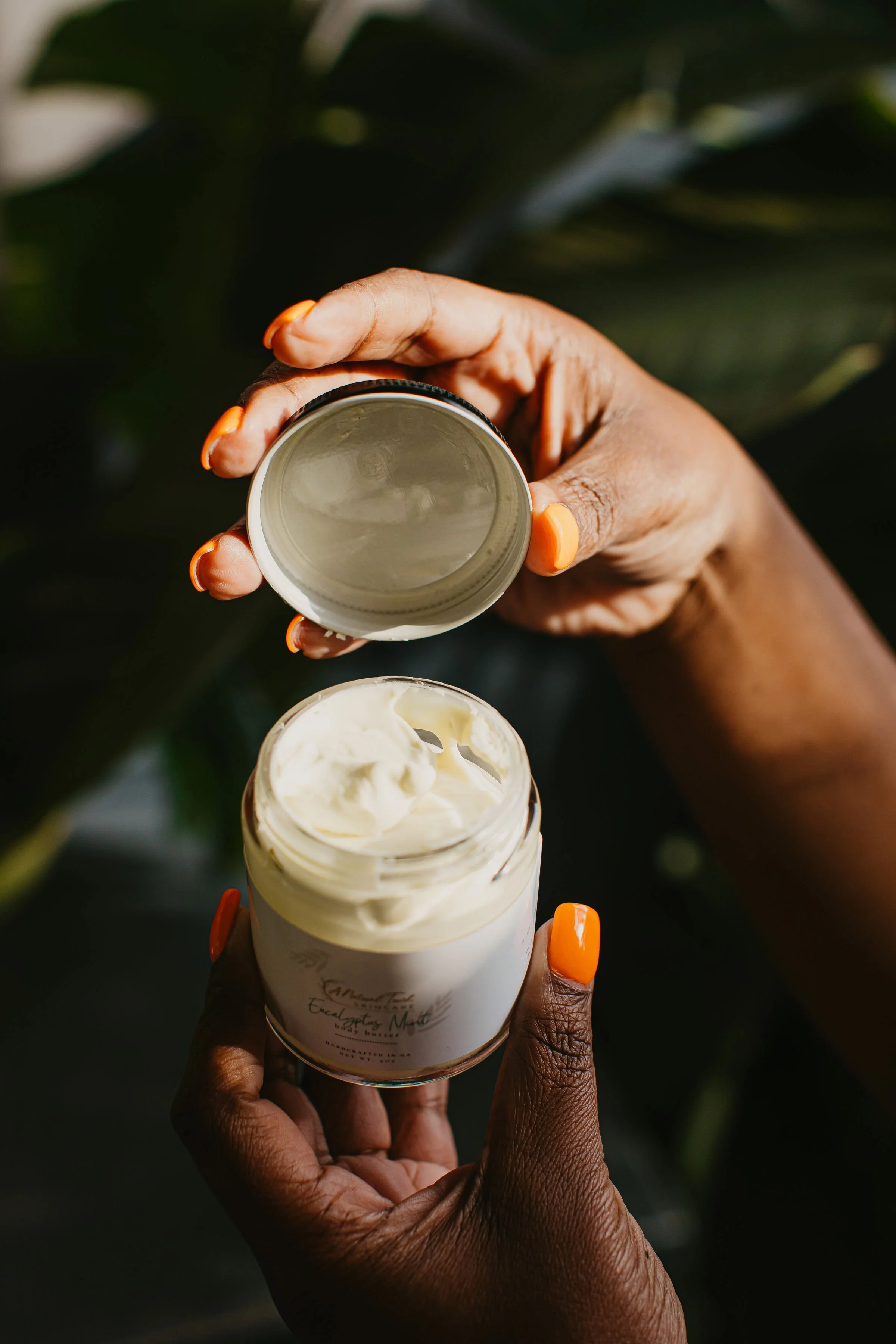 A person with dark skin and painted nails holds a small jar of cream and a lid. The background has green foliage.