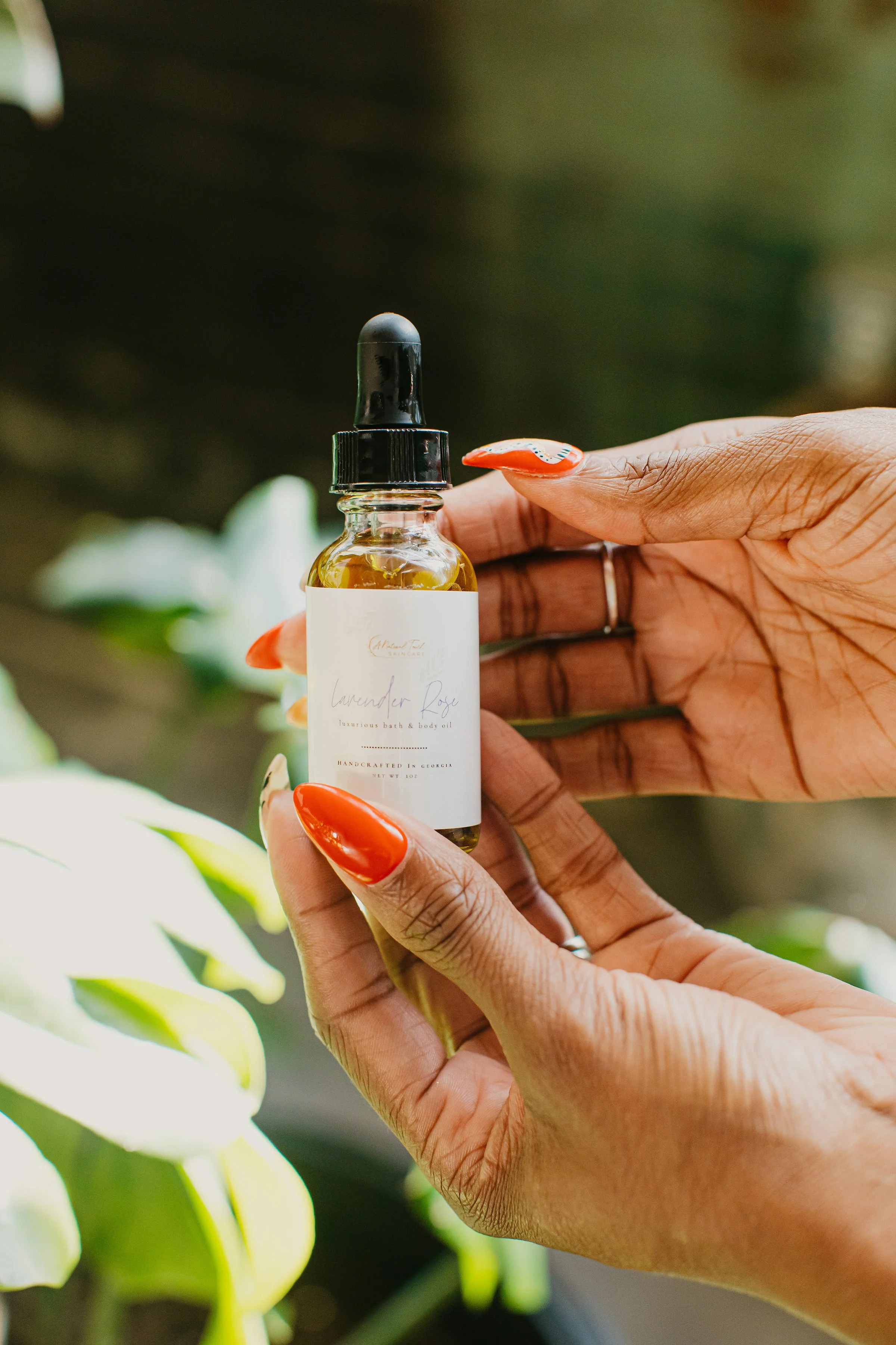 Close-up of a person's hands holding a small bottle of lavender rose body oil with a black dropper lid, with green plants in the background.