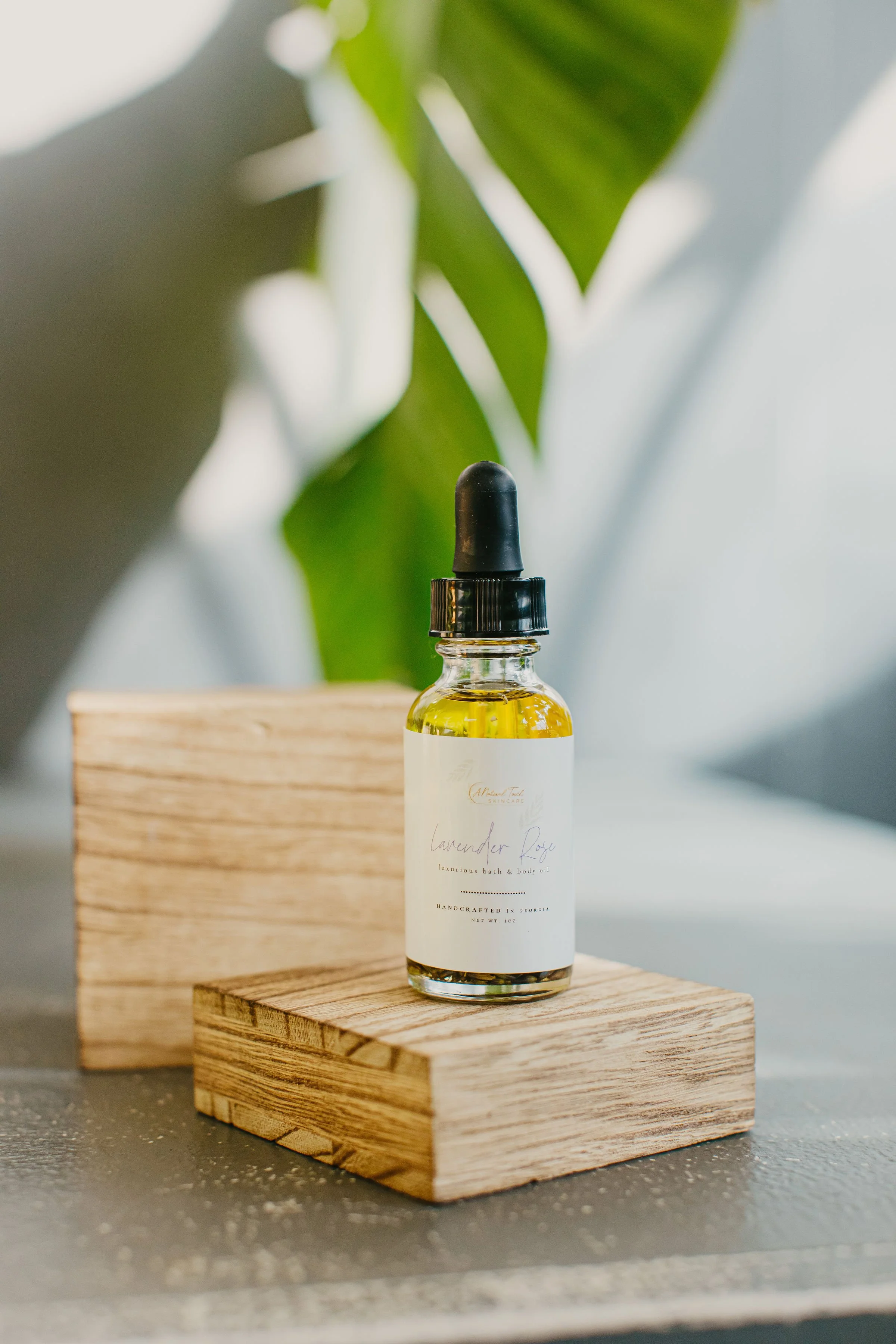 A bottle of lavender rose luxurious bath and body oil with a black dropper cap placed on a wooden block on a gray surface, with a blurred background of green leaves.