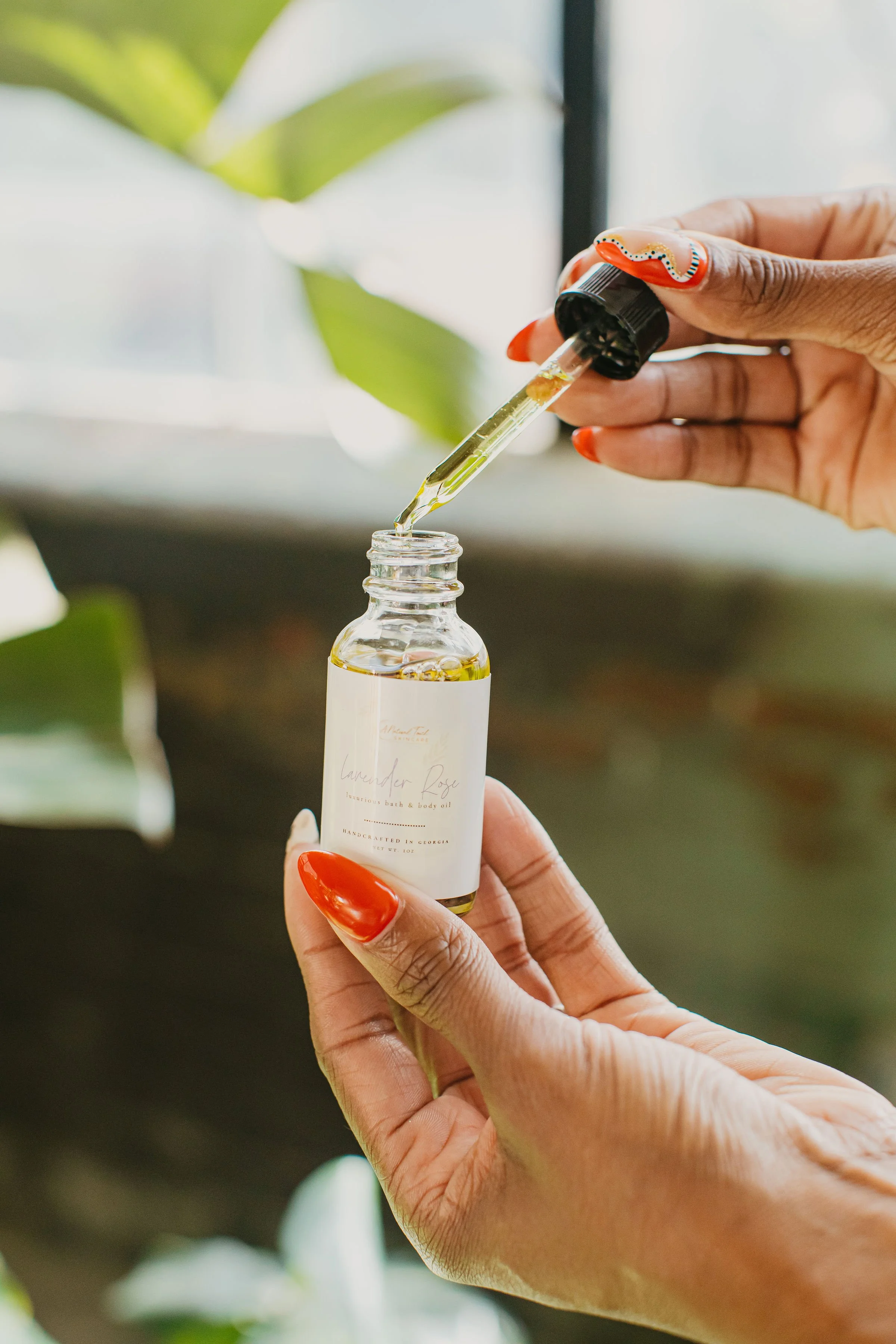 Close-up of a person with painted nails holding a small glass bottle labeled lavender rose bath and body oil, with a dropper releasing oil into the bottle.