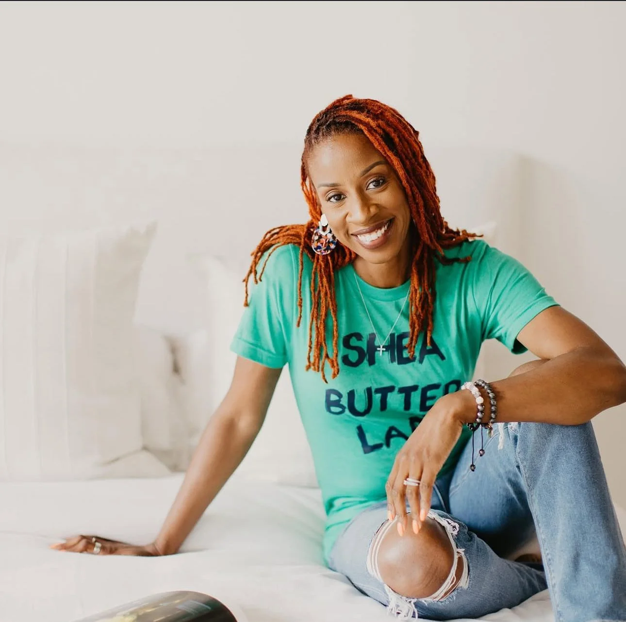 A woman with red dreadlocks smiling at the camera, sitting on a white bed wearing a light blue T-shirt with the words 'SHEA BUTTER LADY' and ripped jeans.