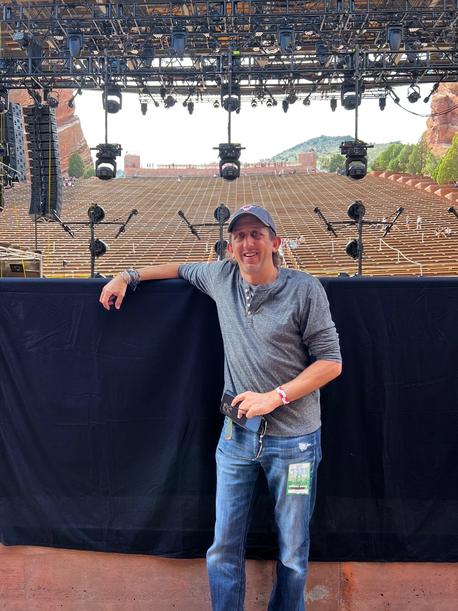 A man standing in front of a stage setup outdoors, with a backdrop of red rocks, trees, and mountains. He is wearing a cap, gray shirt, and jeans, holding a smartphone, and smiling at the camera.
