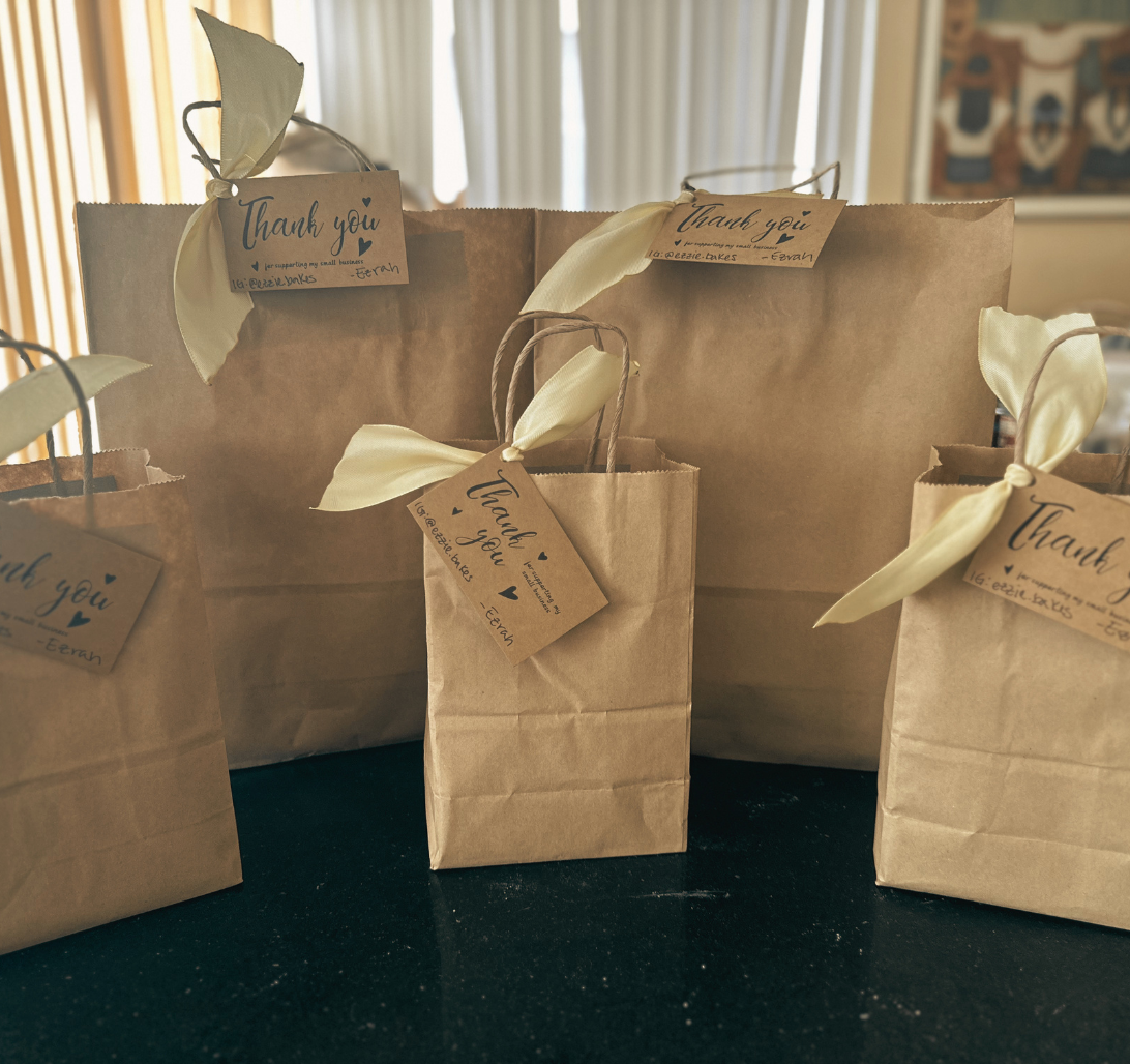 Brown paper gift bags with cream-colored ribbon bows and thank you tags, placed on a black surface with a window in the background.