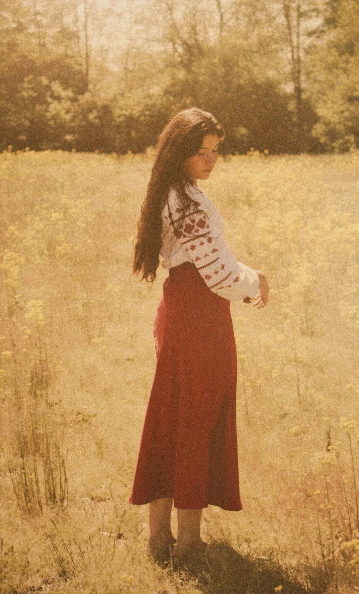 A woman with long dark hair standing in a grassy field with trees in the background, wearing a long red skirt and a white sweater with red patterns.
