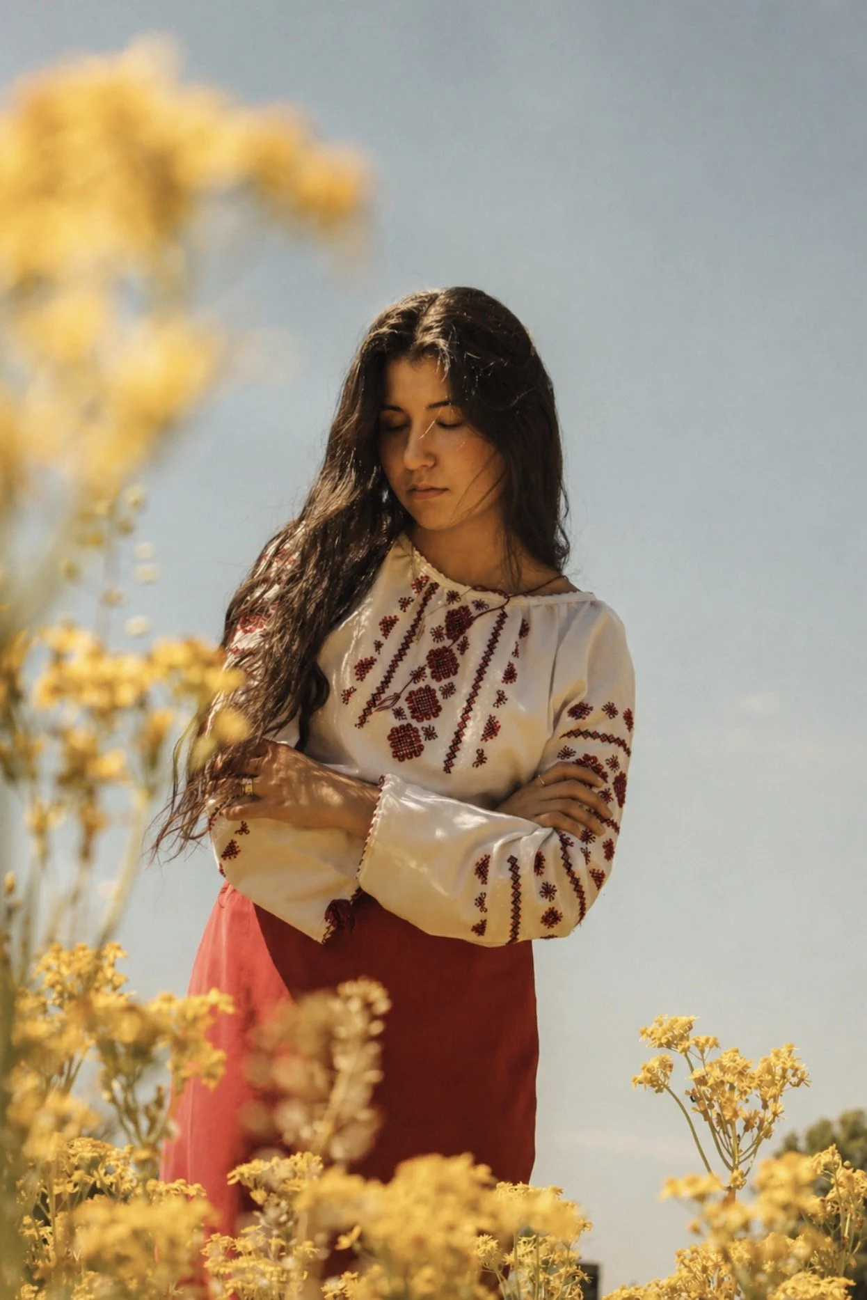 A woman with long dark hair, closed eyes, wearing a white embroidered blouse with red patterns, stands in a yellow flower field against a blue sky background.