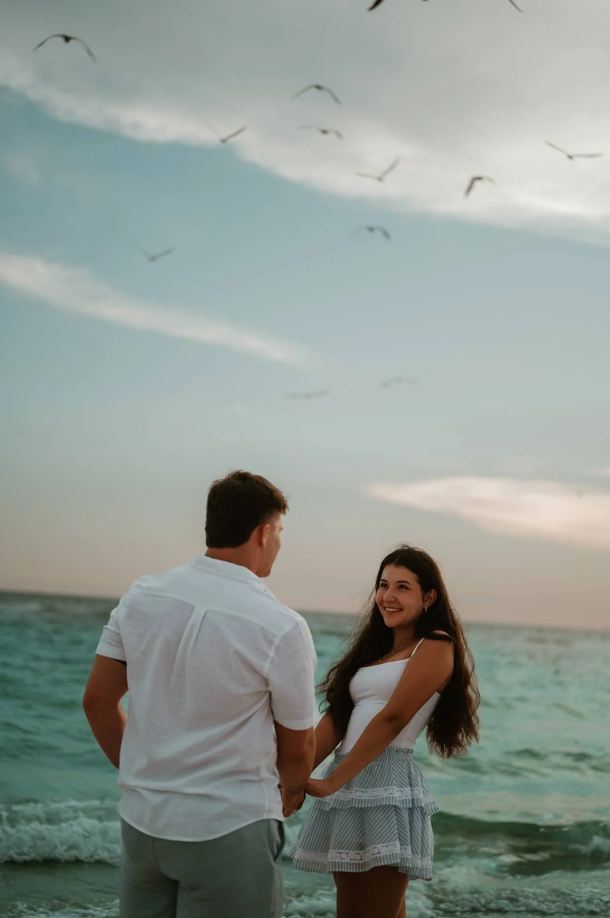 A man and woman holding hands on the beach during sunset, with seagulls flying overhead and the ocean in the background.