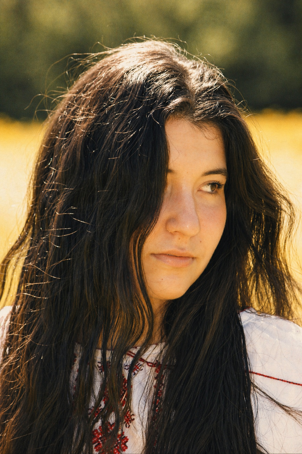 A woman with long, dark, wavy hair outdoors, wearing a white shirt with red embroidery, in a sunny field.