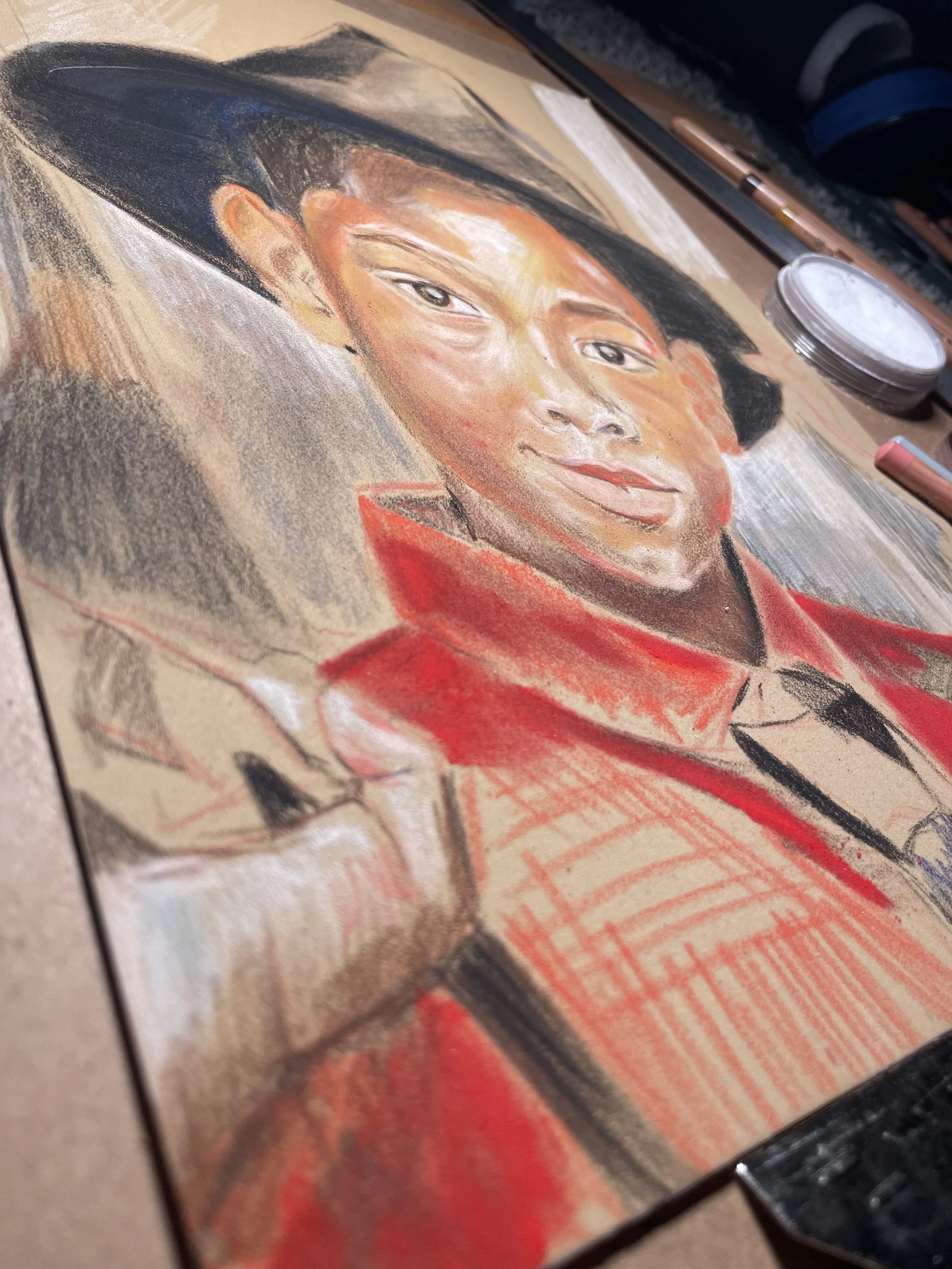 A partially completed portrait of a young child with black hat, resting his head on the hat. drawn with pastels on paper, surrounded by art supplies.