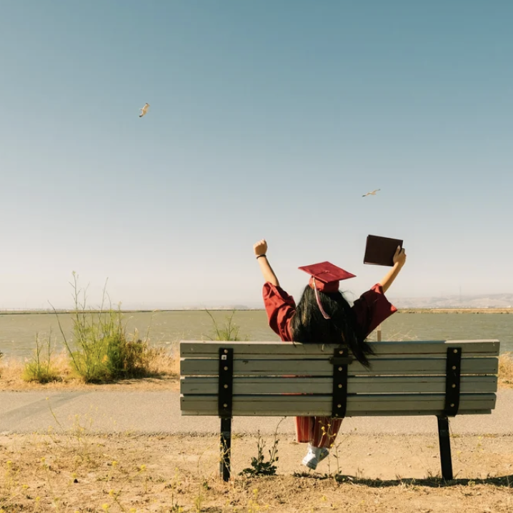 A person in a red graduation gown and cap, sitting on a bench by a body of water, holding a booklet in one hand and raising the other in celebration, with two birds flying in the sky above.