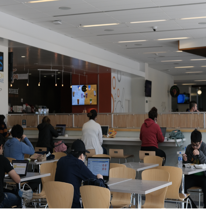 People using laptops and studying in a modern cafe or library with a counter and digital menu screens.