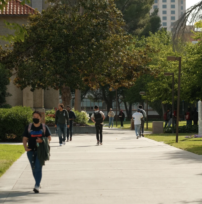 People walking on a university campus or park walkway, surrounded by trees and greenery during daytime.