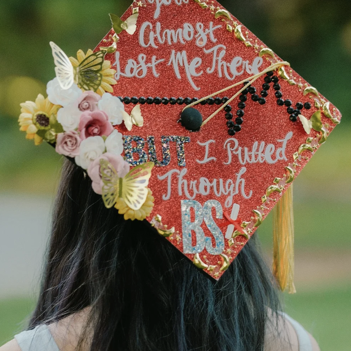 Close-up of a decorated graduation cap with a floral arrangement, butterflies, and colorful text that reads, "Almost Lost Me There, But I Pulled Through BCS".