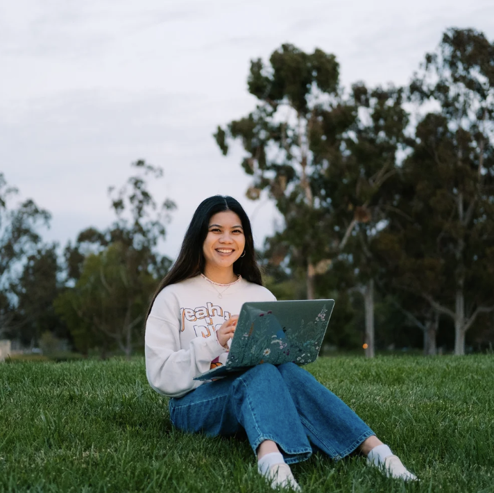 Young woman sitting on grass with a laptop, smiling, outdoors in a park with trees in the background during daytime.