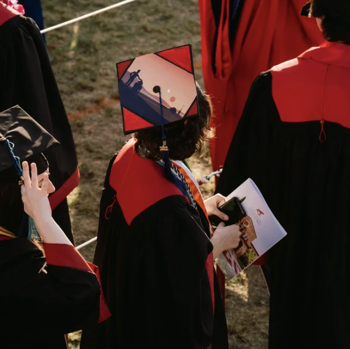 A person in a graduation gown holding a cellphone and a program booklet, with a decorated graduation cap featuring an orange and blue design, during an outdoor graduation ceremony.