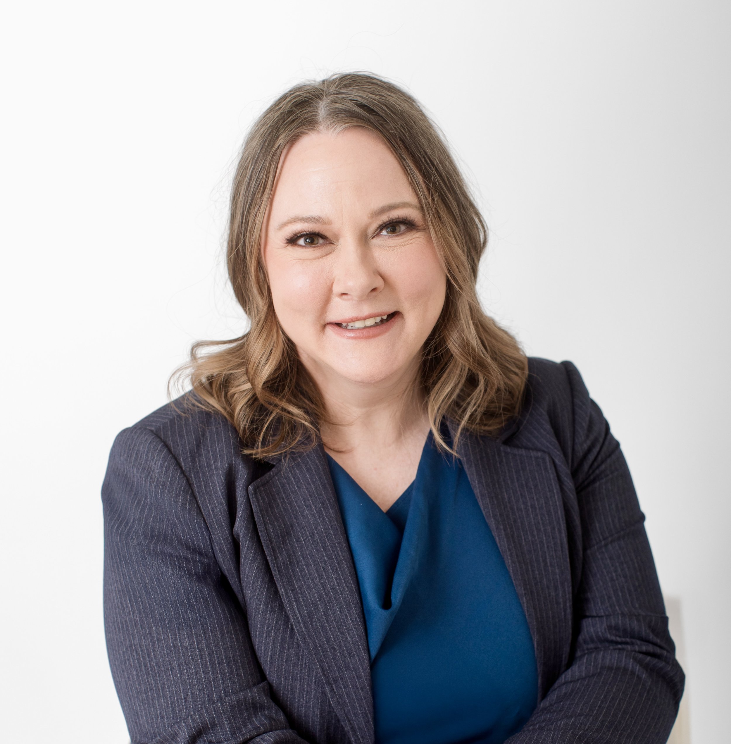 A woman with light brown, wavy hair wearing a navy blazer and dark blue top, smiling against a plain white background.