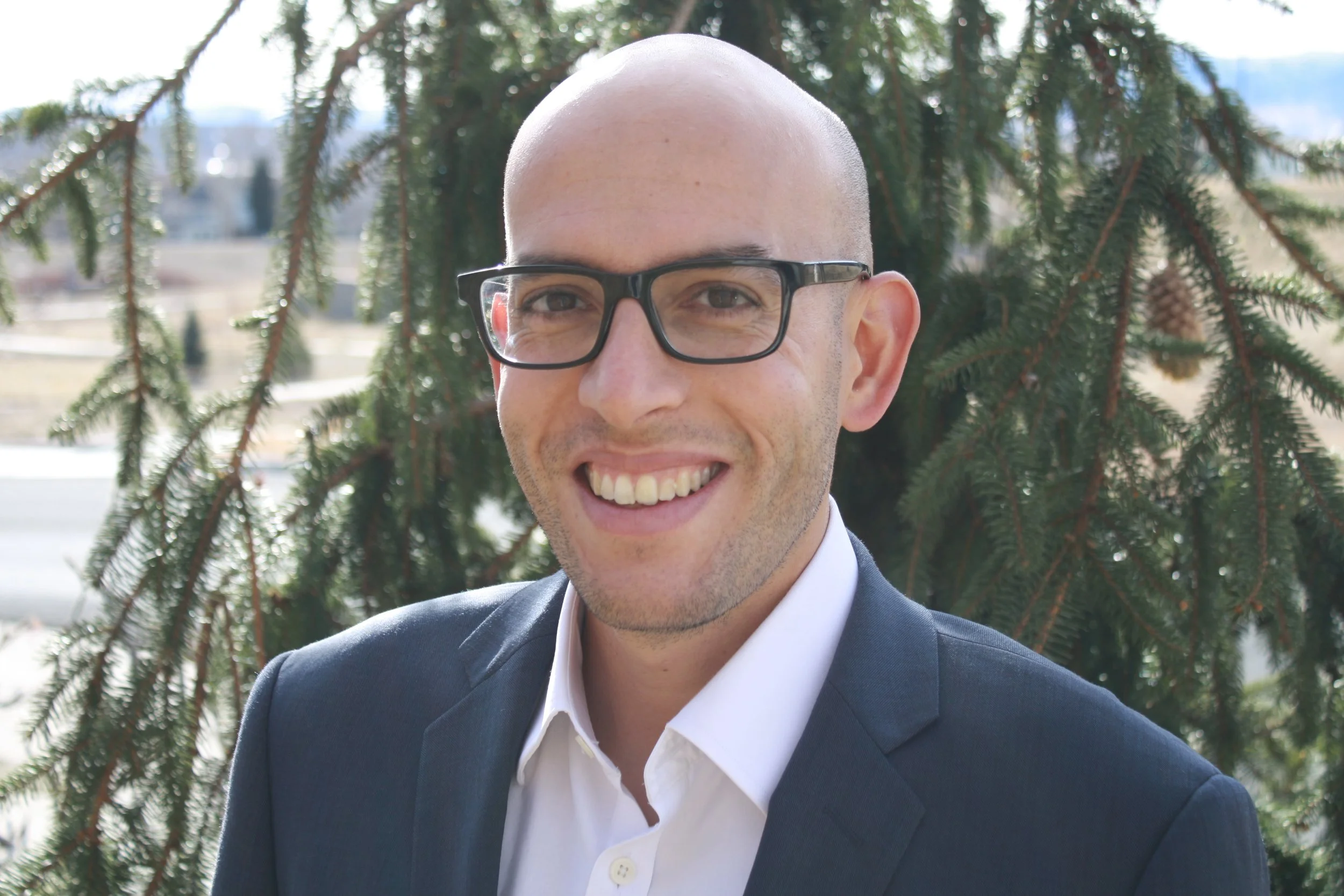 A smiling man in a suit and glasses standing outdoors in front of a tree.