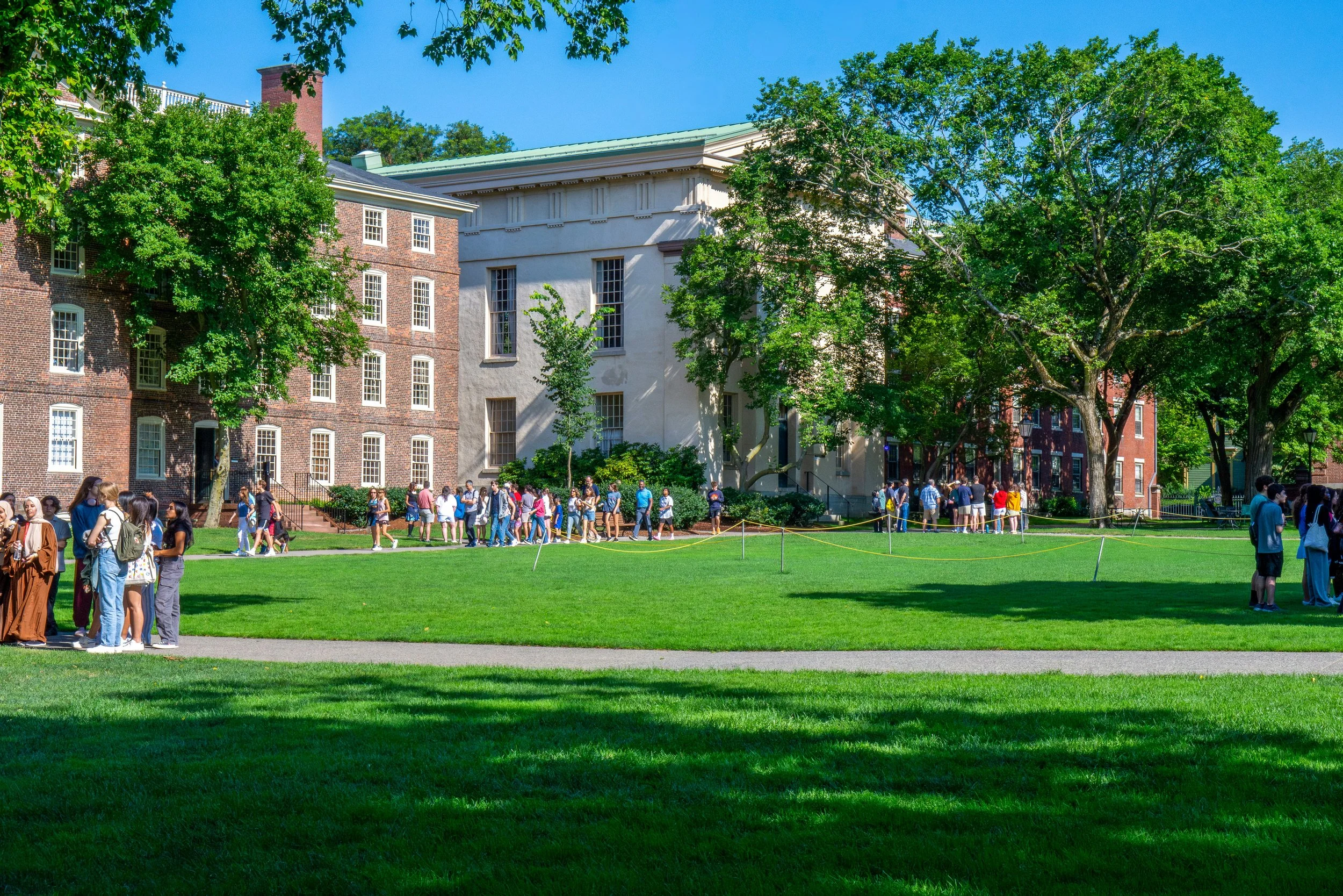 Students lining up outside a historic university building with trees and a green lawn on a sunny day.
