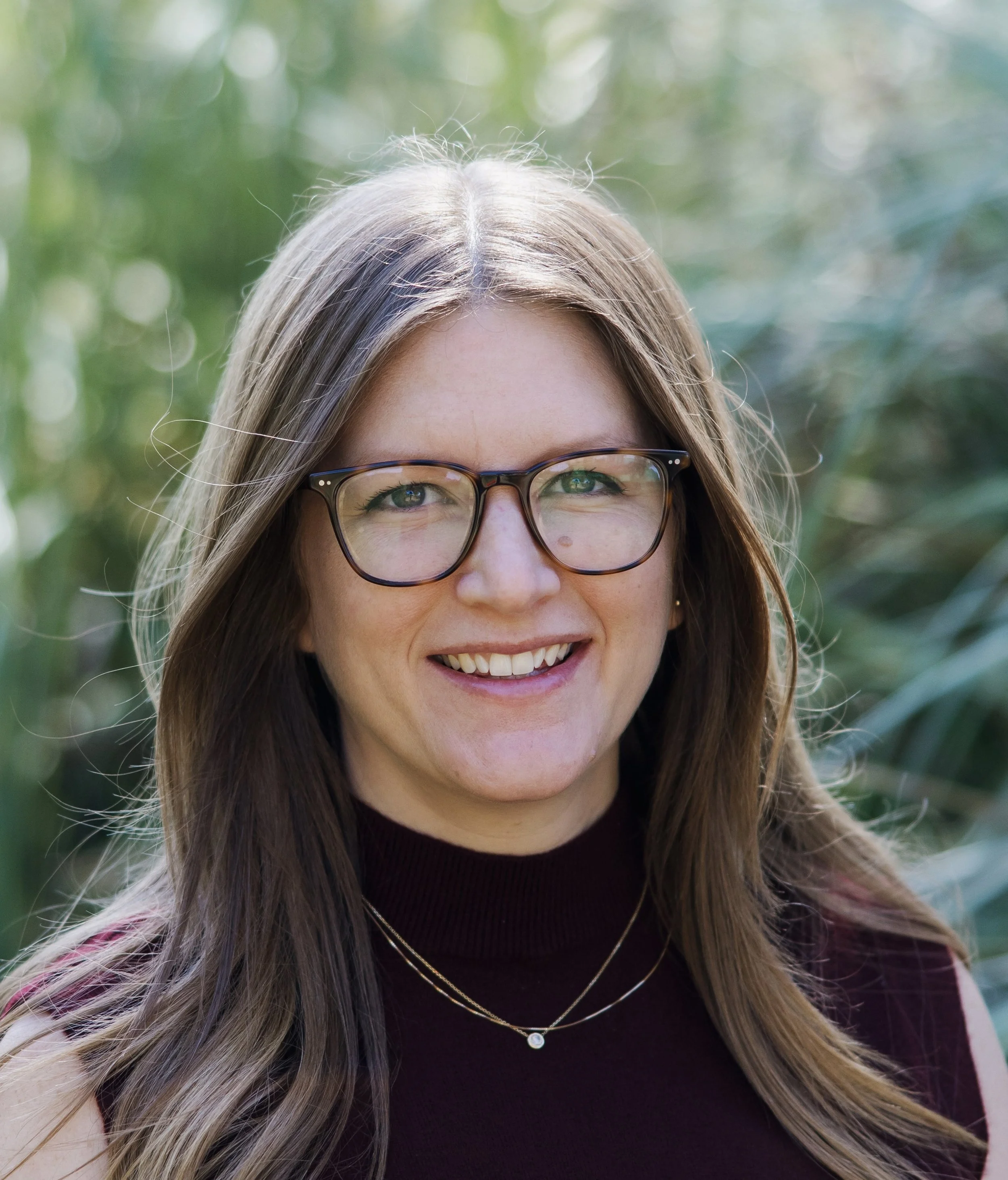 Close-up of a woman with long brown hair, wearing glasses, a black turtleneck, and layered necklaces, smiling outdoors with blurred greenery background.