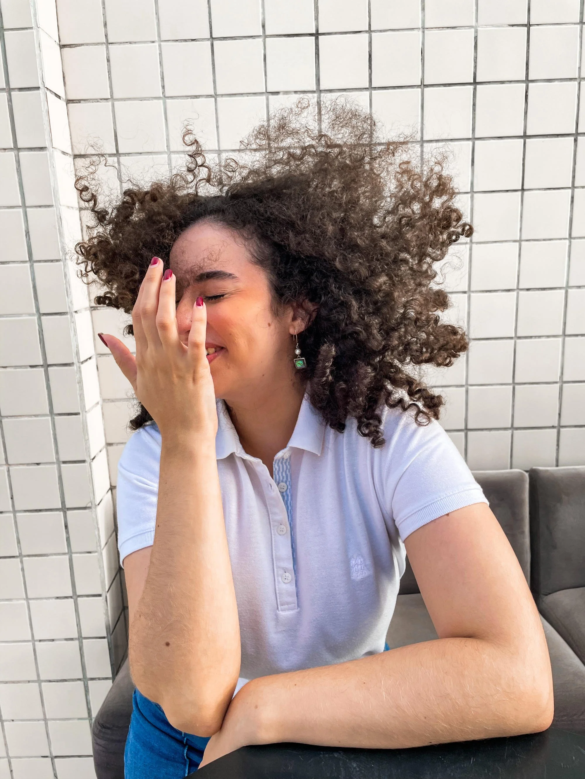 A woman with curly hair and earrings sitting against a tiled wall, covering part of her face with her hand, smiling with closed eyes.