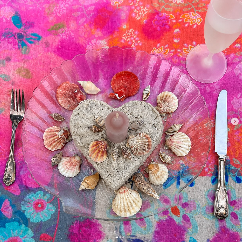 A decorative table setting featuring a pink glass plate with a heart-shaped concrete centerpiece, a pink candle, and scattered seashells; a fork on the left, a knife on the right, and a glass of champagne on the top right, all on a colorful floral tablecloth.
