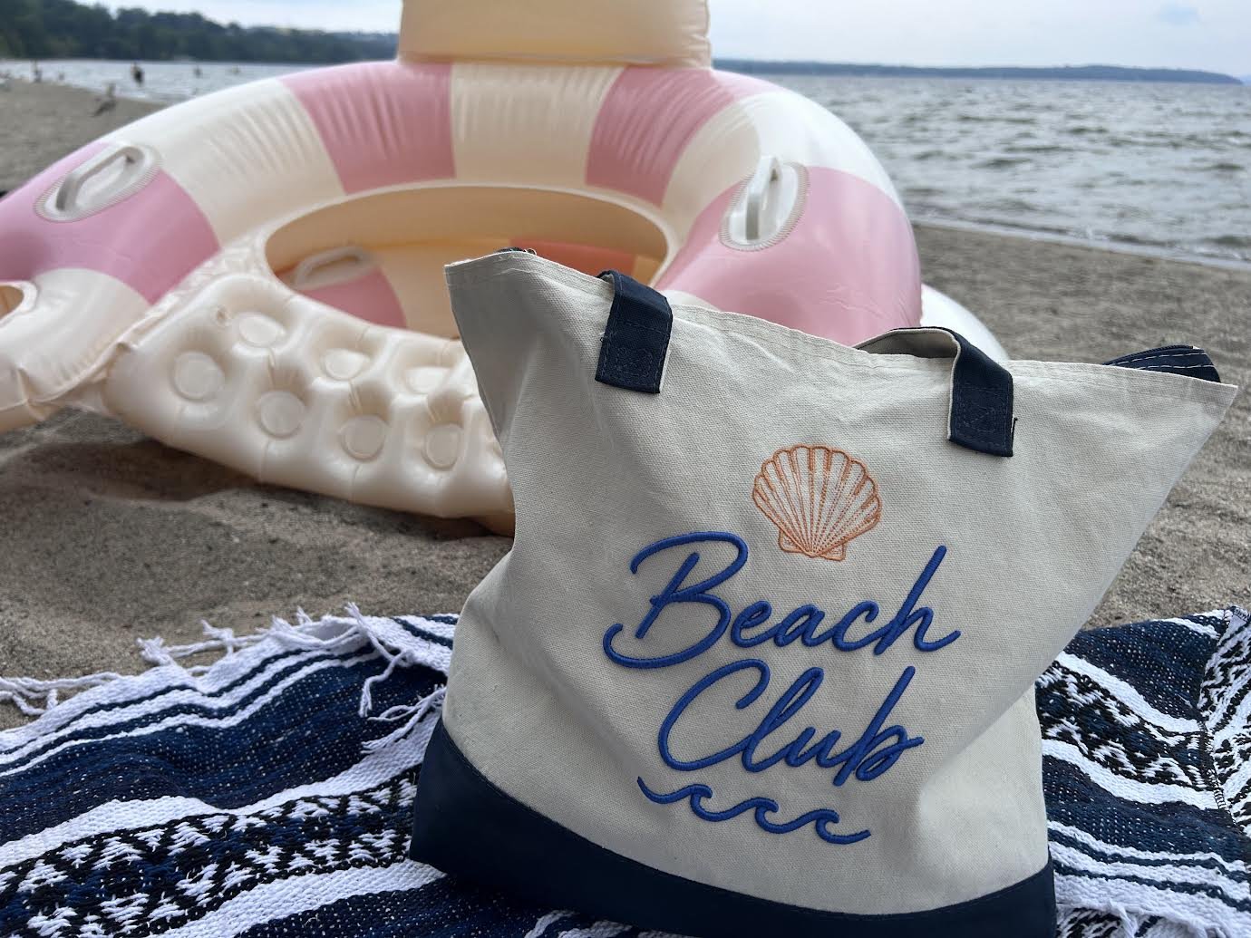 A beach scene with a pink and white inflatable ring and a tote bag with a seashell and the words "Beach Club" on it, placed on a striped beach towel near the water.
