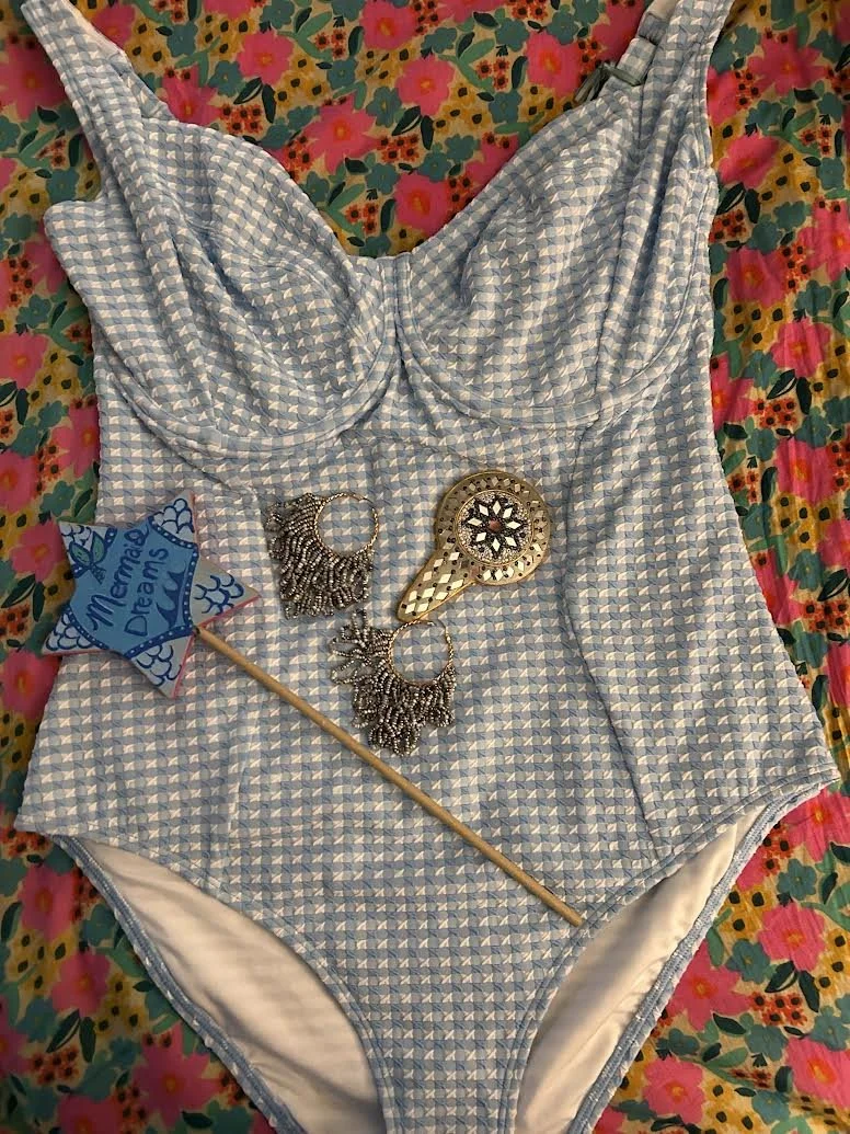A blue and white checkered vintage-style swimsuit is laid flat on a floral blanket. On top of the swimsuit, there is a small sign that says "Memories of Dreams" attached to a stick, a pair of ornate silver earrings, and a handheld mirror with a decorative handle.