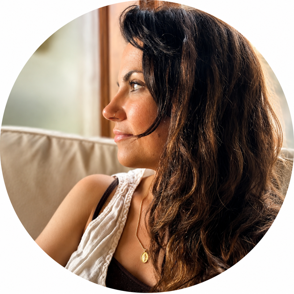 Side profile of a woman with wavy brown hair and a nose piercing, sitting on a beige couch, wearing a gold necklace with a pendant, and looking to the left.