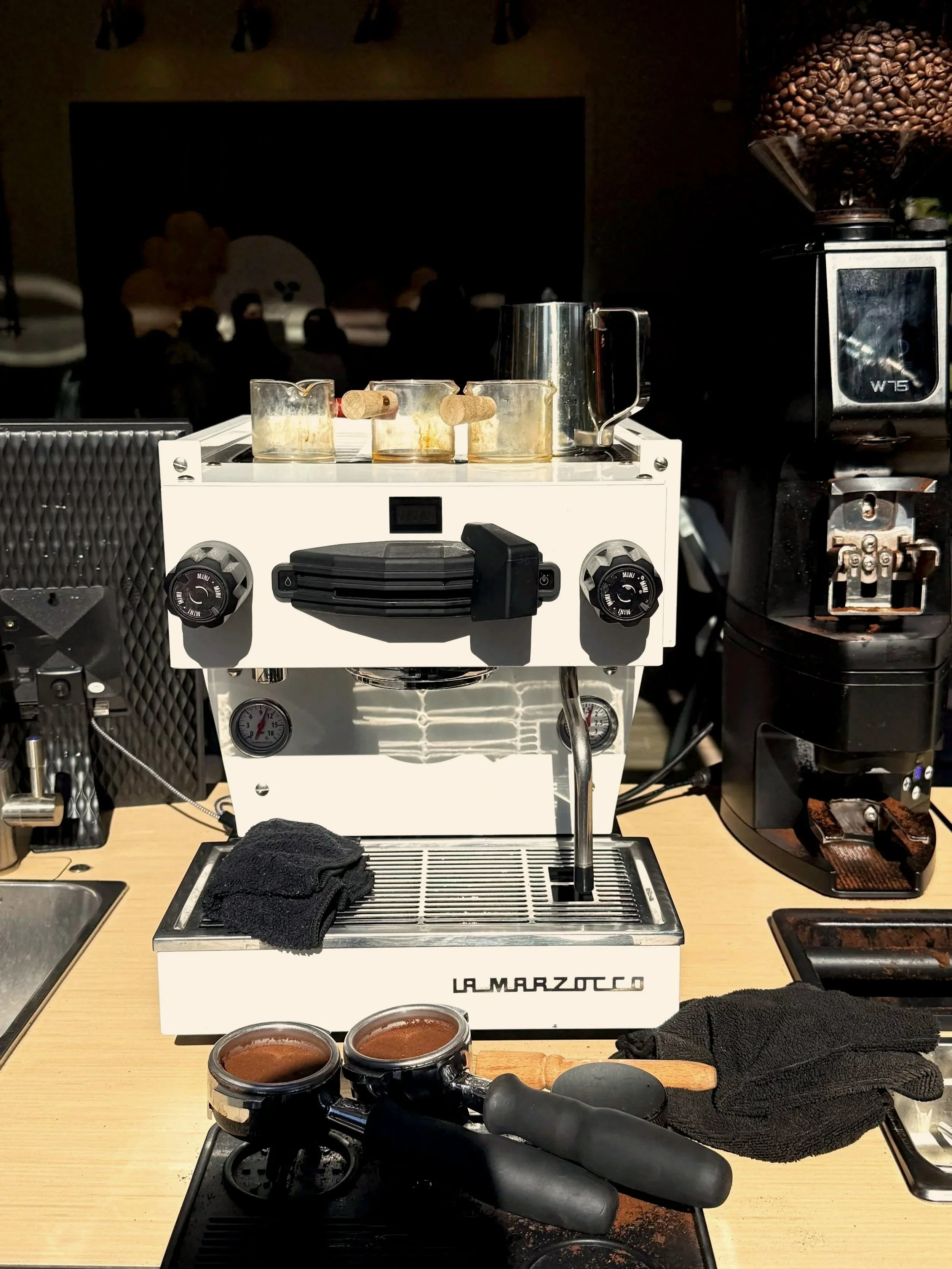 A La Marzocco espresso machine on a wooden counter, with a portafilter filled with coffee grounds in front, and a black towel beside it. Behind the machine, there is a grinder filled with coffee beans and a black textured surface. The scene suggests a coffee preparation setup in a café or barista workspace.