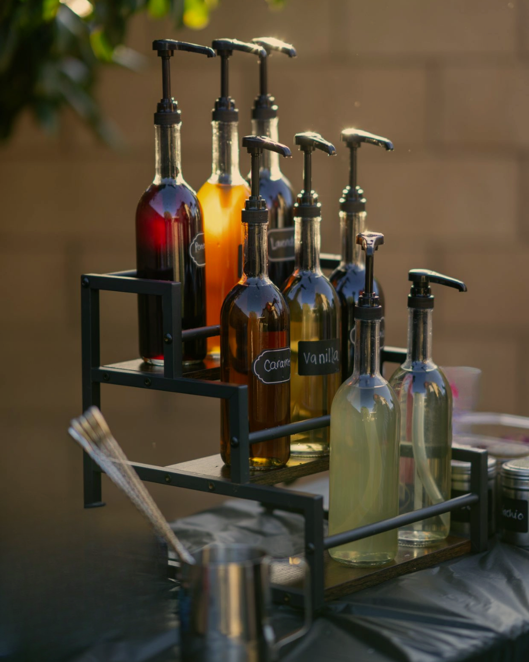 Assorted flavored syrups in glass bottles with labels for caramel, vanilla, and other flavors, arranged on a two-tiered black metal and wood stand.