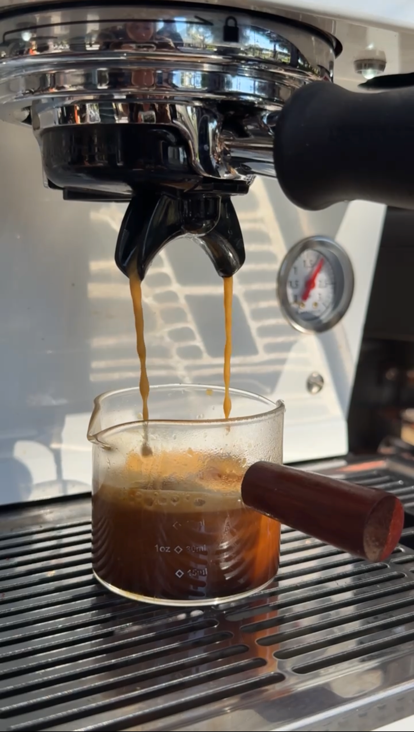 Coffee flowing from a coffee machine into a glass cup with a wooden handle, with a pressure gauge visible on the machine.