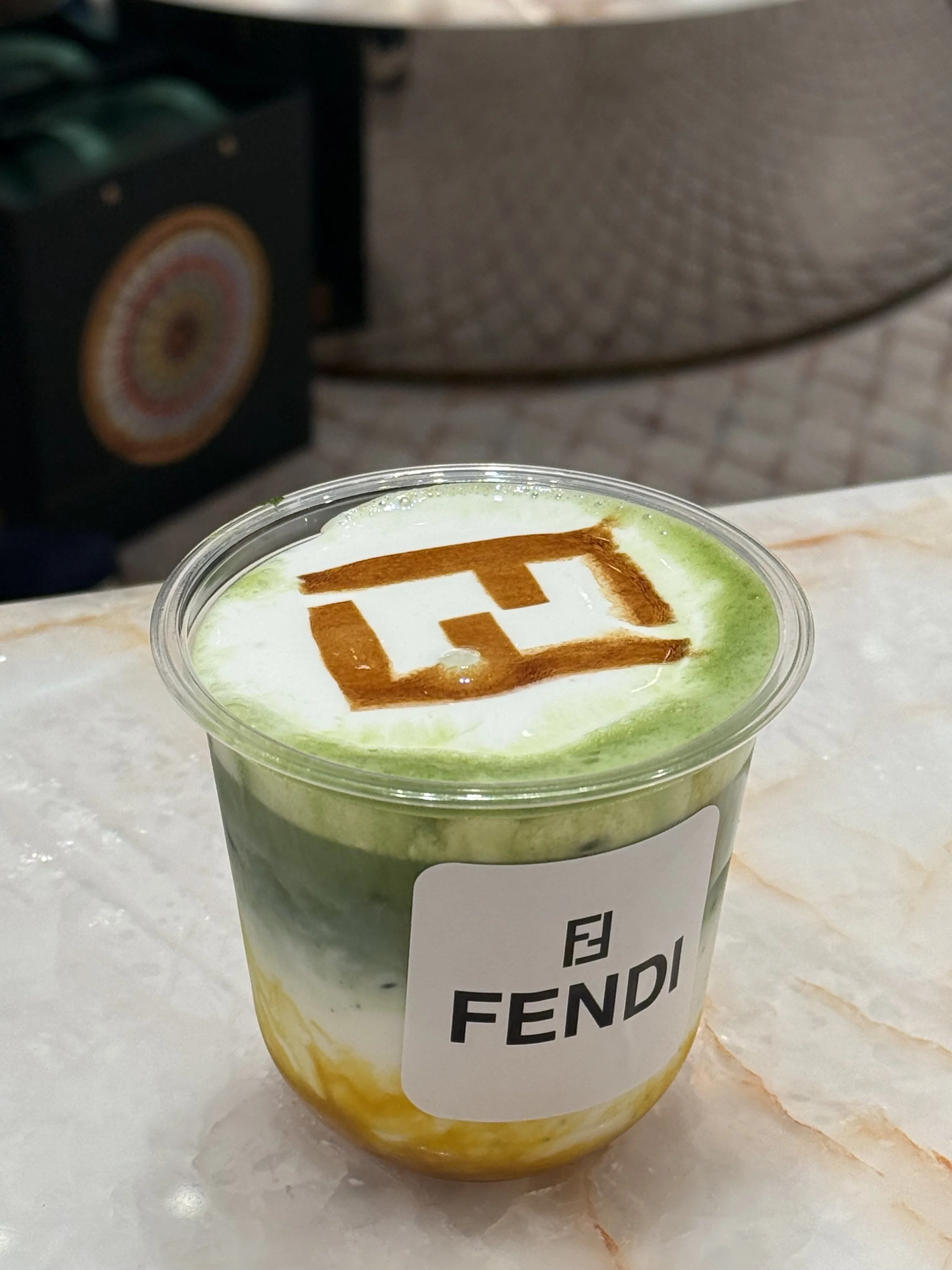 A coffee station at an outdoor event with cups, a La Marzocco espresso machine, and coffee beans in a grinder, with blurred people in the background.