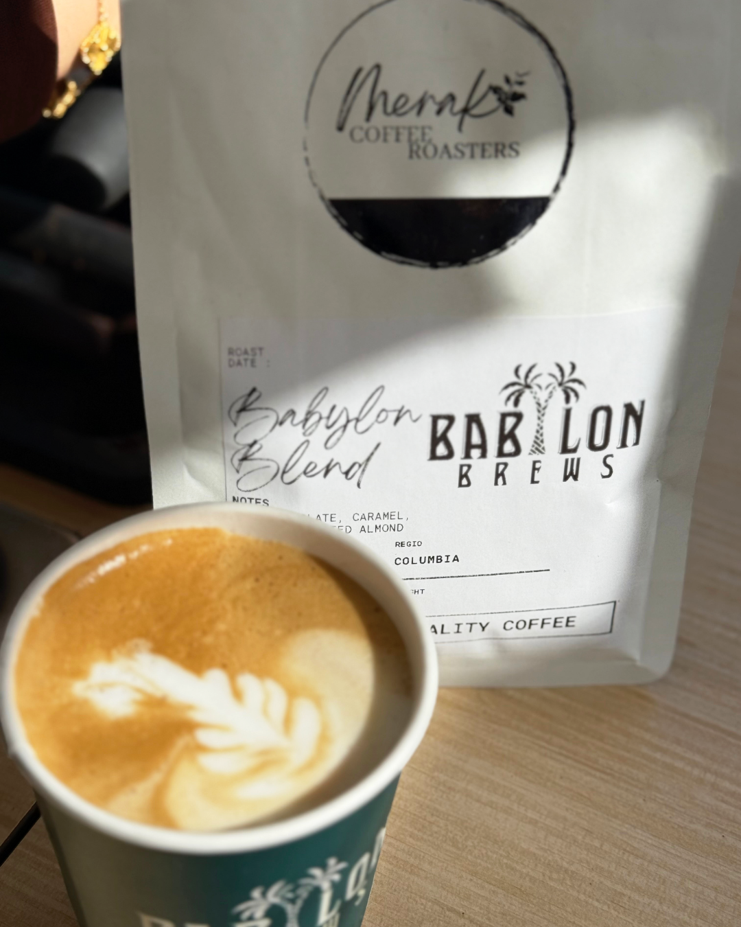 A cup of coffee with latte art next to a bag of Babylon Blend coffee beans from Bab Lor Brews on a wooden table.