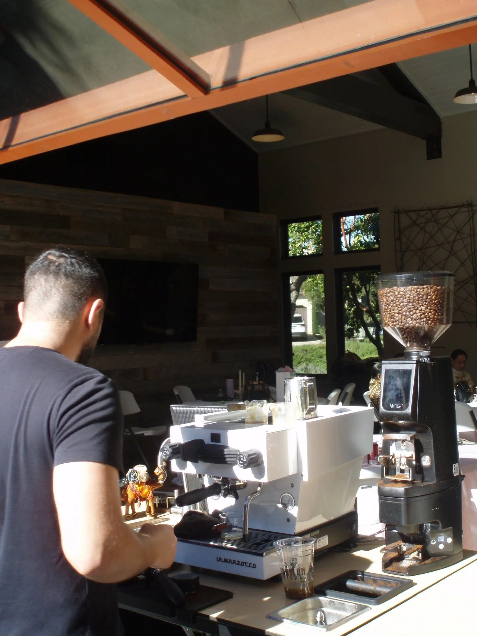 A man with short dark hair wearing a black shirt operating a coffee machine on a white counter in a modern cafe, with a large container of coffee beans on a grinder and a glass of iced coffee nearby.