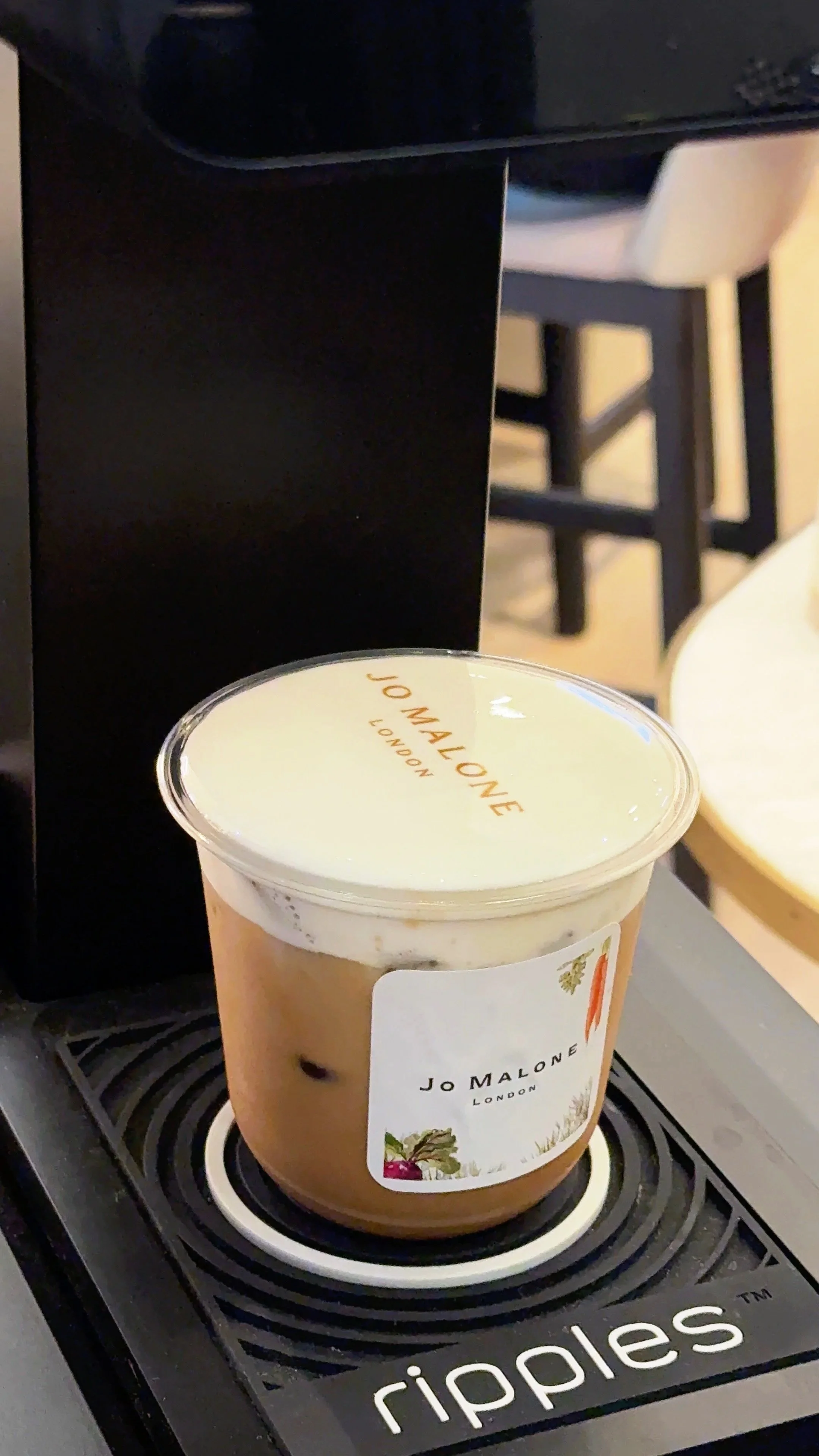 A hand holding a clear plastic cup of iced coffee with a transparent lid, featuring a blank white circular label with the text "Your Logo Here" in red cursive font. The background shows green foliage.