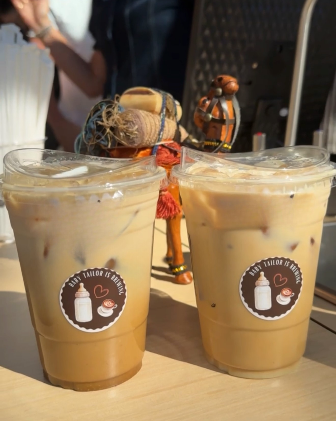 Two iced coffee drinks in plastic cups with a logo that says 'Baby Tailor is Brewing' on a wooden table, with a decorative figurine of a person in the background.
