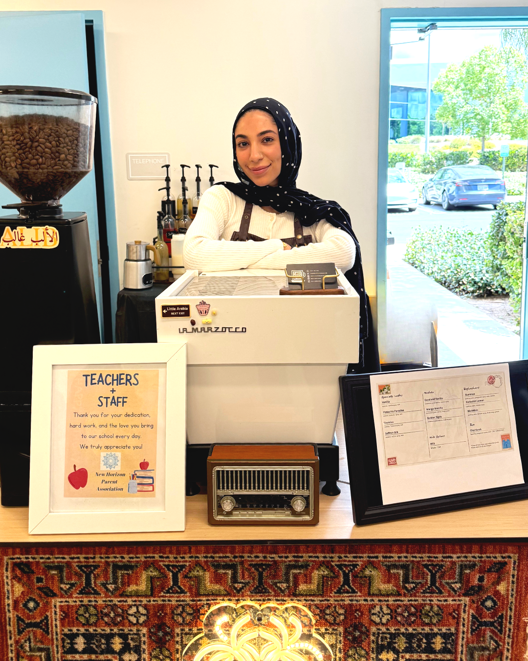A smiling woman wearing a hijab and apron stands behind a small coffee stand with a cash register in a bright, indoor space near a large window showing parked cars and trees outside.