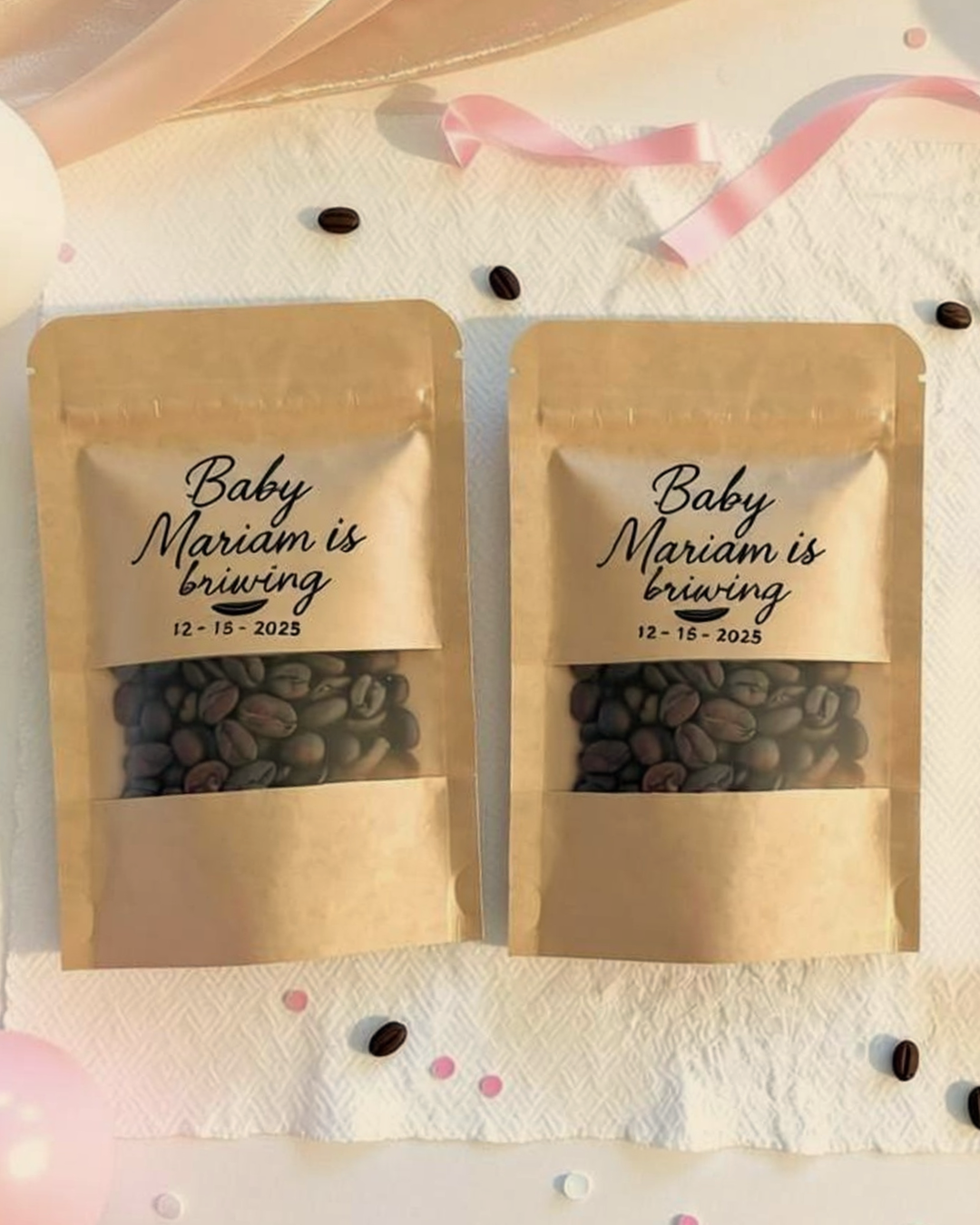 Two beige resealable bags of coffee beans with labels reading "Baby Mariam is brewing" and the date 12-15-2025, surrounded by scattered coffee beans, pink ribbons, and pink and white confetti on a white textured surface.