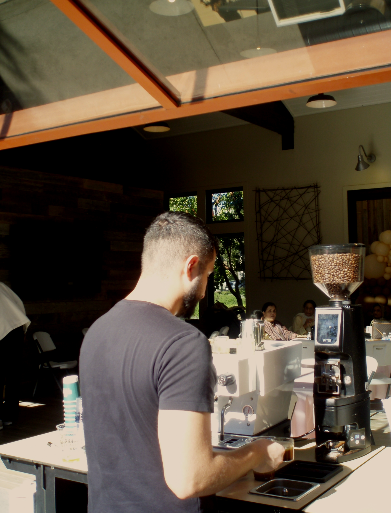 A man with a short beard and dark hair, wearing a black t-shirt, operating a coffee machine at a coffee stand in a bright indoor space with large windows showing greenery outside. Several people seated in the background inside the cafe.