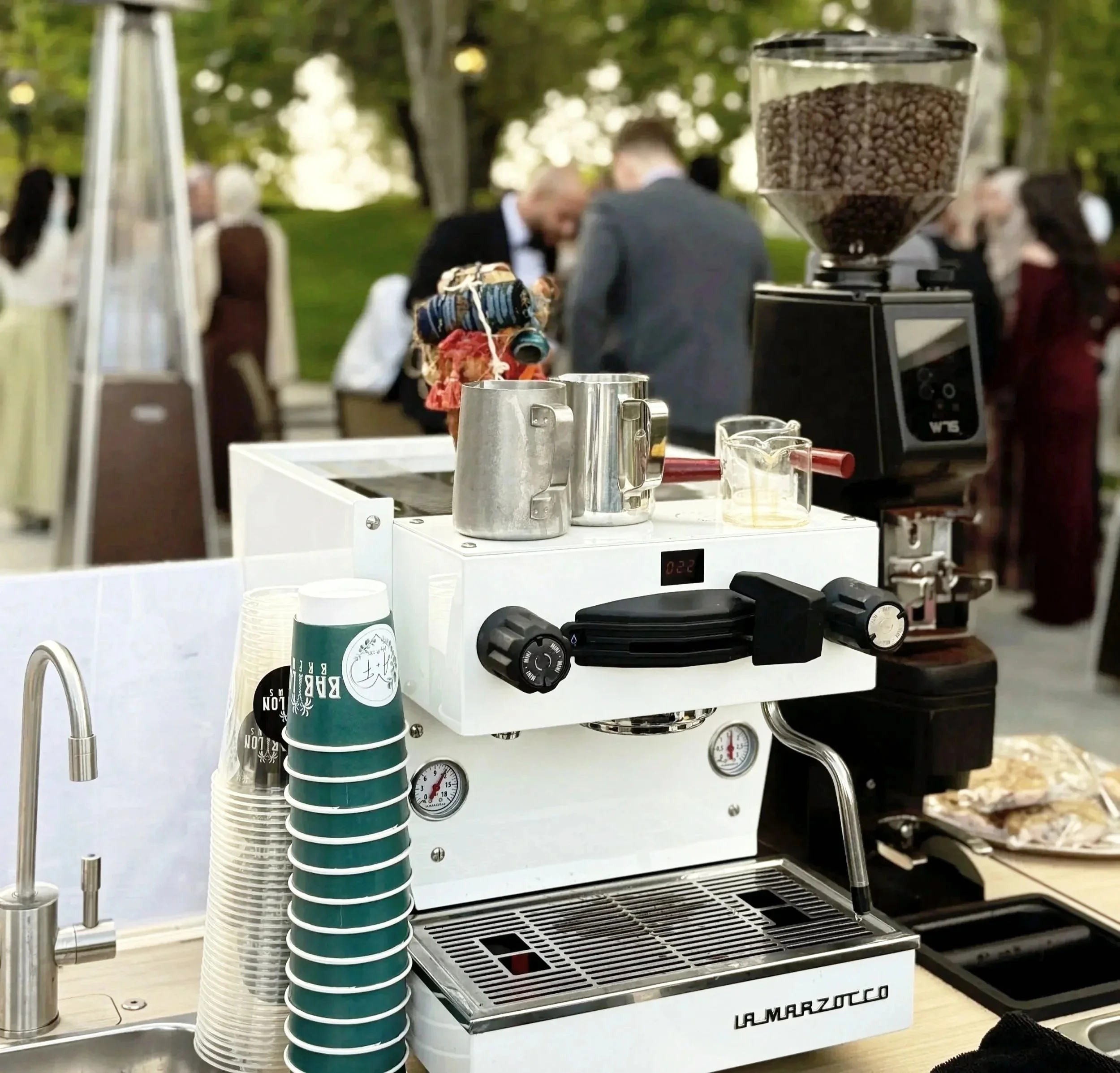 A mobile coffee bar with a sign that reads "Love is Brewing" in a decorated event venue with string lights and tables set for dining.