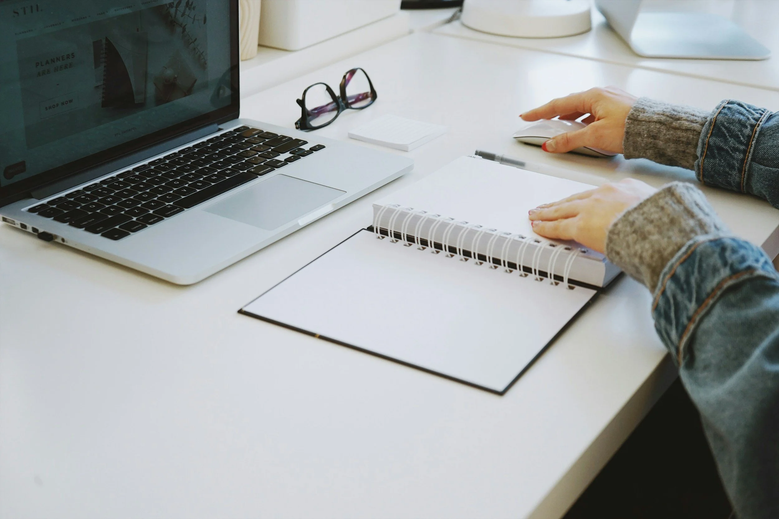 Person in a gray sweater using a mouse at a white desk with a MacBook, reading glasses, a notepad, and a pen.