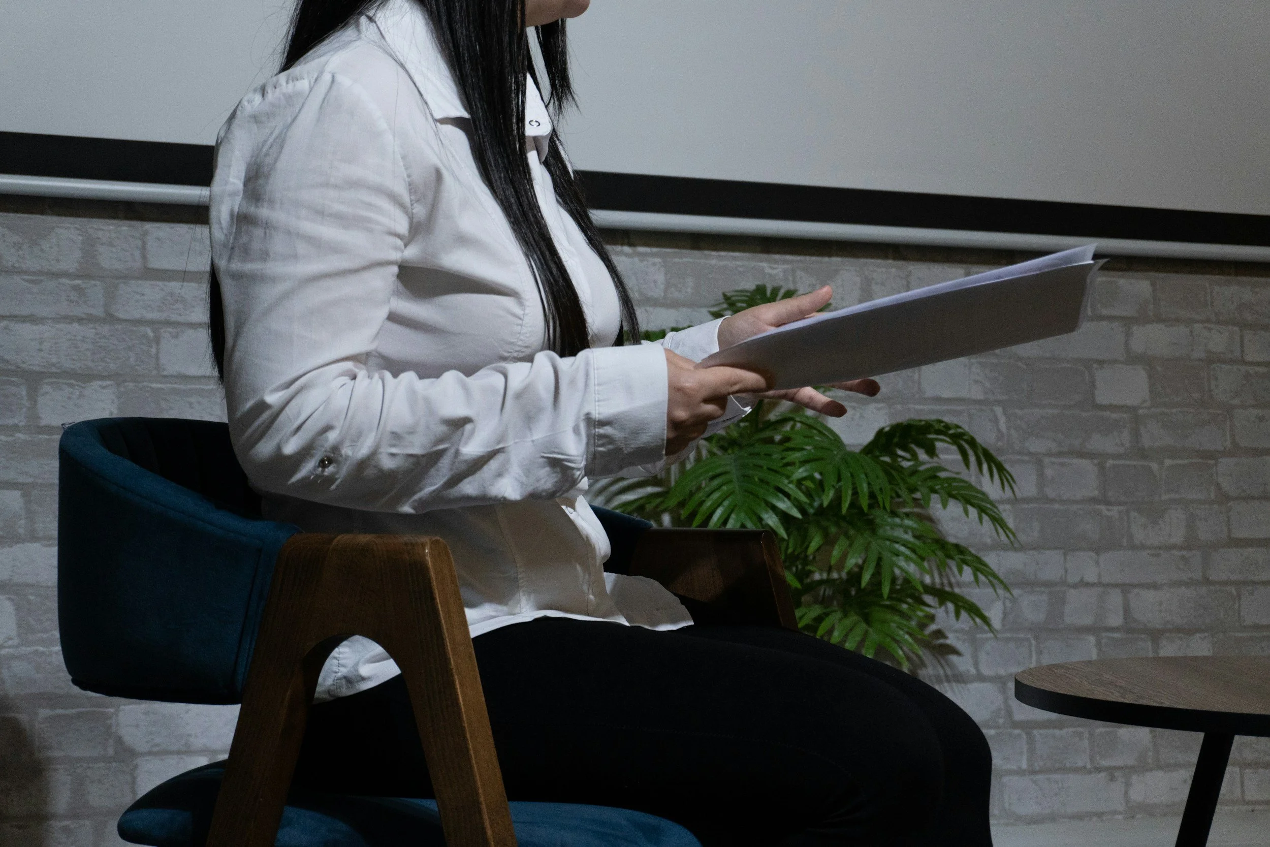 A person holding a beige fabric sample over an open book on a wooden table, with ceramic dishes and textured stone samples also on the table.