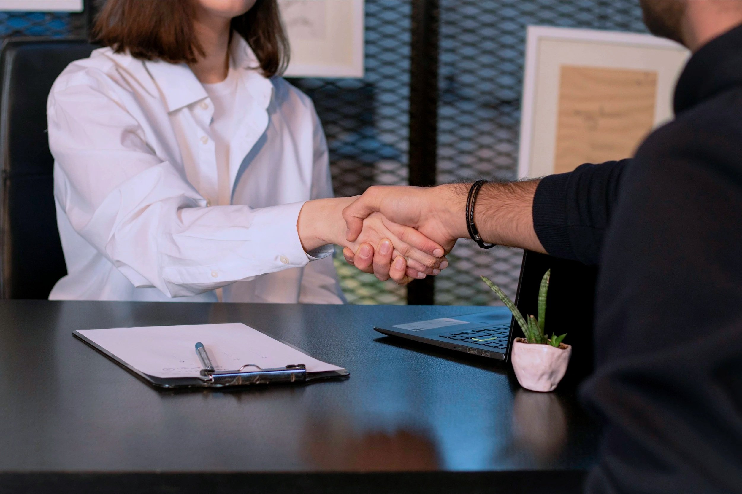 Close-up of a handshake between a woman with shoulder-length brown hair wearing a white button-up shirt and a man with short dark hair wearing a black long-sleeved shirt, seated at a desk with a laptop, notepad, pen, and small potted plant.