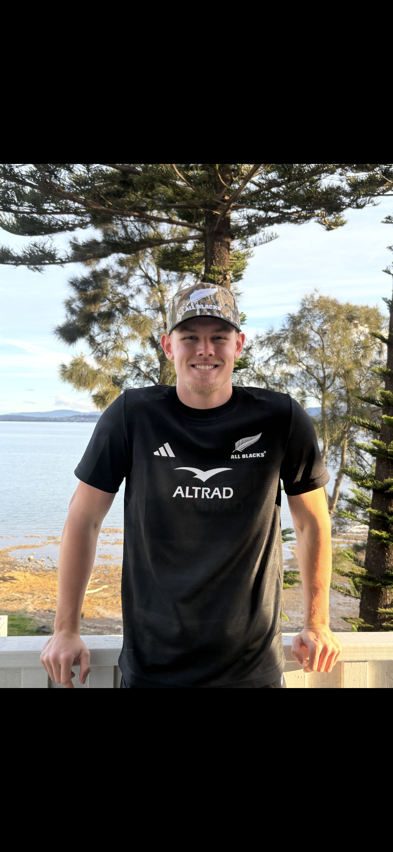 Young man standing outdoors near water, smiling, wearing a black sports jersey and a camouflage cap, with trees and a body of water in the background.